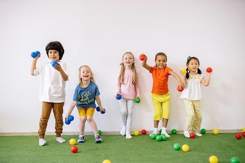 5 Kinder stehen mit dem R&uuml;ckend vor einer Wand  und spielen mit kleinen B&auml;llen. Alle Kinder halten B&auml;lle in der Hand und viele weitere B&auml;lle in der verschiedenen Farben liegen auf dem Boden. 