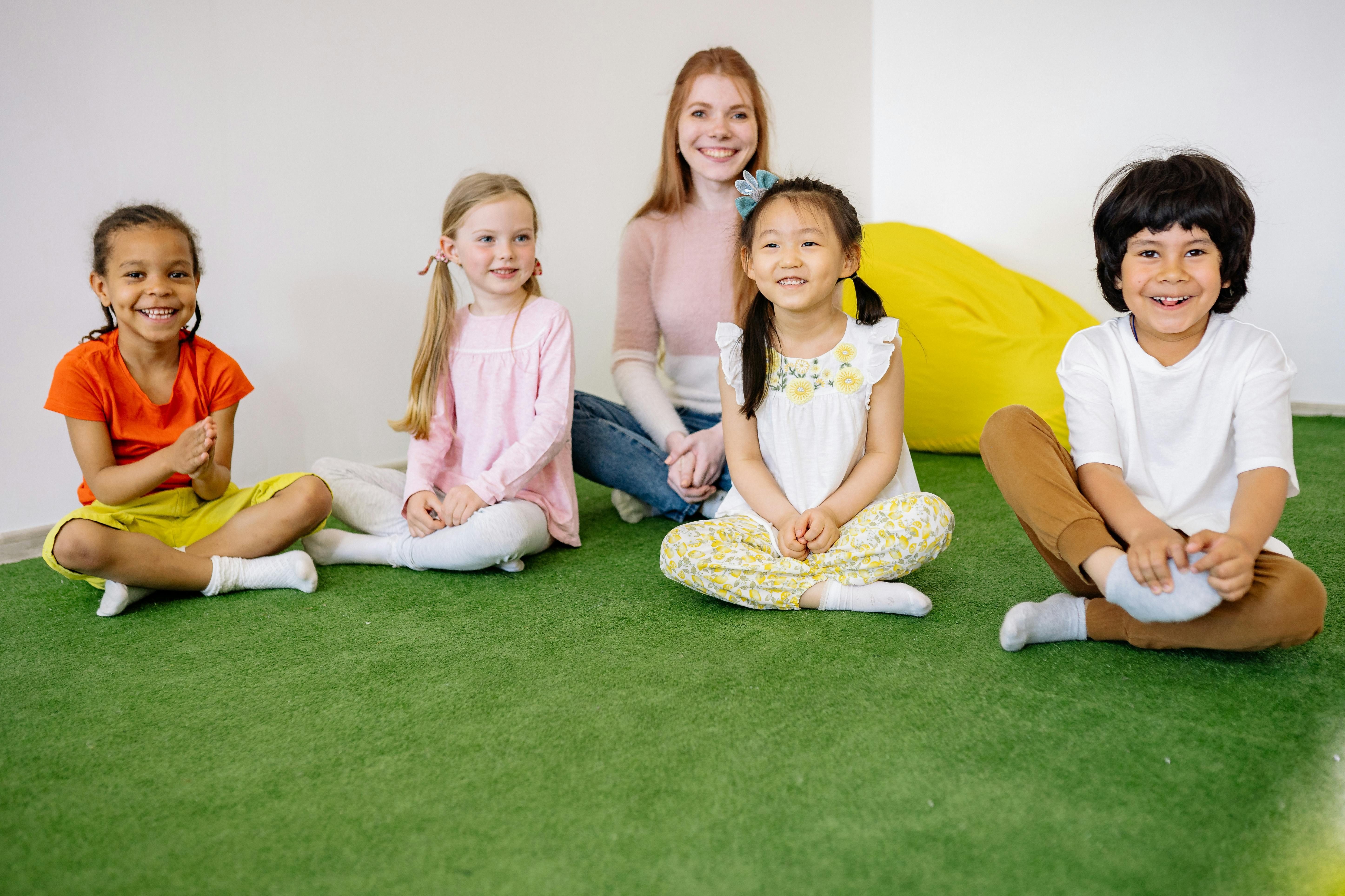 4 Kinder sitzen in einem leeren Raum im Schneidersitz vor einer Frau, die ebenfalls im Schneidersitz sitzt und alle lachen oder l&auml;cheln. Hinter ihnen liegt ein gelber Sitzsack.