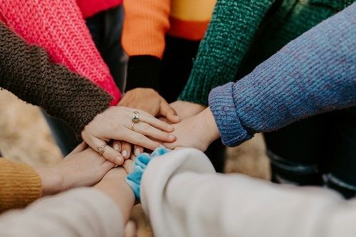 Mehrere Personen stehen in einem Kreis. Von ihnen sind haupts&auml;chlich die Arme und H&auml;nde zu erkennen. Die H&auml;nde werden alle &uuml;bereinander in der Mitte zusammengef&uuml;hrt.  