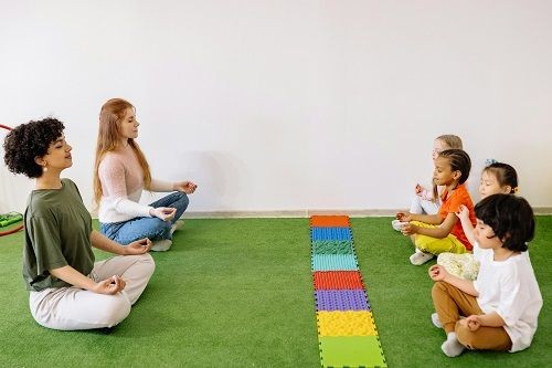 Zwei Frauen sitzen 4 Kindern mit etwas Abstand gegen&uuml;ber. Alle sitzen im Schneidersitz mit den H&auml;nden auf den Knien in einer Meditationsposition, Zwischen ihnen liegt eine Reihe aus aneinander gebauten bunten Puzzleteilen. 