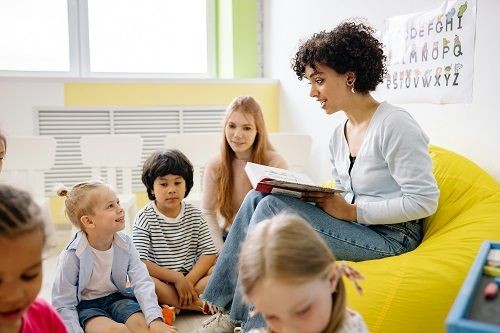 Zwei Frauen und mehrere Kinder sitzen in einem Raum. Eine der Frauen liest den Kindern ein Buch vor. 