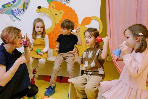 Eine Frau sitzt in einem bunten Raum mit 4 Kindern und alle halten eine Glocke in der Hand und heben diese teils an ihr Ohr. 