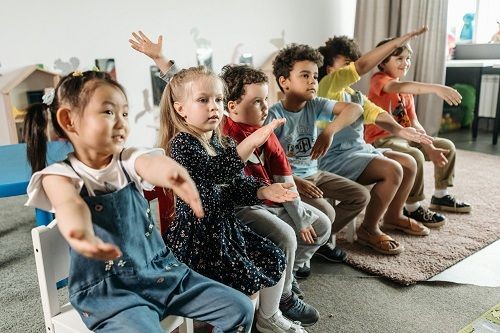 Kinder sitzen in einem Raum in einer Reihe nebeneinander und geben verschiedene Handzeichen. 