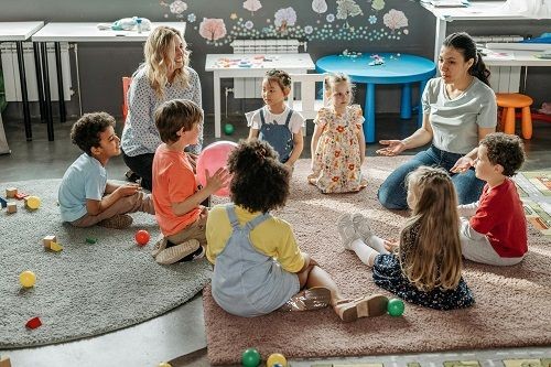 Mehrere Kinder sitzen mit zwei Frauen in einem Kreis und unterhalten sich. Um sie herum stehen Kindertische und es liegt Spielzeug auf dem Boden. 