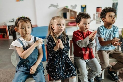 Kinder sitzen in einer Reihe auf St&uuml;hlen nebeneinander. Einige der Kinder klatschen in die H&auml;nde. 