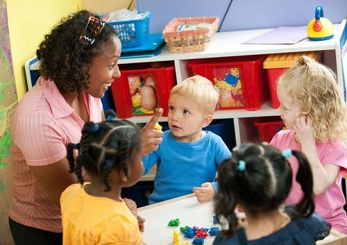 Eine Frau sitzt mit mehreren Kindern an einem kleinen Tisch. Sie h&auml;lt den Zeigefinger nach oben und l&auml;chelt. Die Kinder schauen Sie an und auf dem Tisch liegt bunte Knete. 