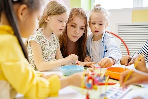 Eine Frau sitzt mit mehreren Kindern an einem kleinen Tisch. Sie schaut den Kindern beim malen mit Wasserfarben zu. 