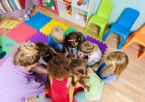 Eine Frau und mehrere Kinder knien auf einem Teppich aus Puzzleteilen zusammen und schauen auf K&auml;rtchen mit Buchstaben darauf. 