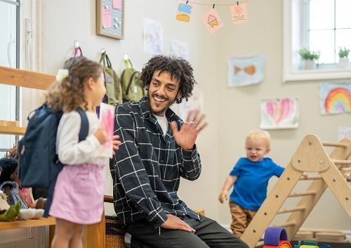 Ein Kind steht in einer Kita und h&auml;lt ein gemaltes Bild vor sich. Ein Mann winkt ihr zu, w&auml;hrend im Hintergrund ein kleineres Kind sich an einem kleinen Kletterger&uuml;st festh&auml;lt. 
