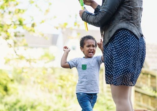 Eine Person, welche nur ab den Schultern zu sehen ist, und eine Flasche mit Seifenblasenmischung in der Hand h&auml;lt und ein Kind stehen im Garten. Das Kind hat die Arme in der Luft und macht ein unzufriedenes oder w&uuml;tendes Gesicht. 