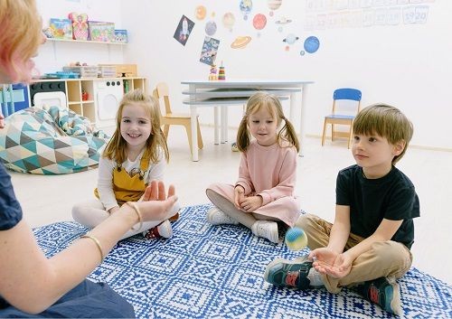 Eine Frau sitzt auf einem Teppich in einem Kitaraum vor 3 Kindern. Eines der Kinder ist kurz davor einen kleinen Ball zu fangen. 