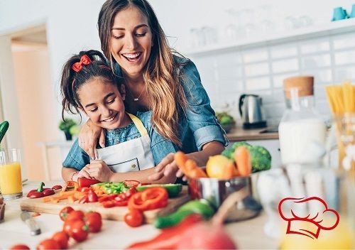 Eine Frau steht hinter einem Kind vor einer Arbeitsfl&auml;che in einer K&uuml;che. Vor ihnen steht ein Topf mit mehreren Gem&uuml;sesorten sowie ein Brett mit geschnittenen Paprika und Karotten.