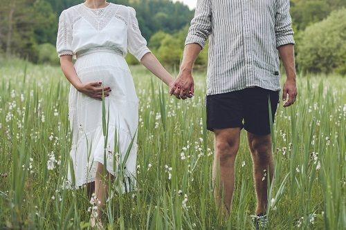 Schwangere Frau im wei&szlig;en Kleid und Mann mit kurzer Hose laufen Hand in Hand &uuml;ber eine bl&uuml;hende Wiese.