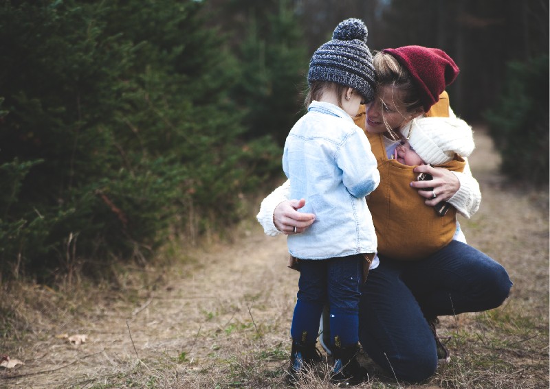 Eine Mutter hat ihr Baby in der Trage und ein Kleinkind neben sich. Sie hat sich hingehockt um mit dem Kleinkind auf Augenh&ouml;he Kontakt zu haben. 