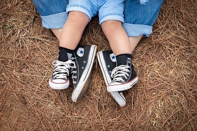 Die F&uuml;&szlig;e einer erwachsene Person mit einem Kind auf dem Scho&szlig;, von dem auch nur die F&uuml;&szlig;e zu sehen sind. Sie sitzen im Heu und tragen beide ein gleiches Modell von schwarzen Turnschuhen.
