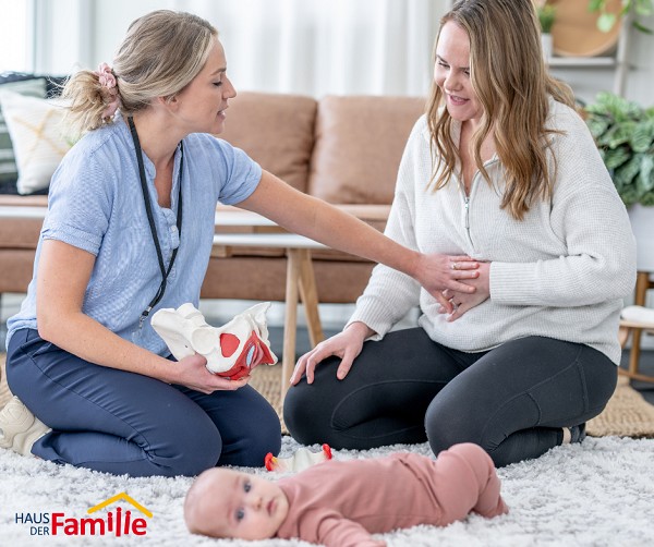 Eine Fachkraft erkl&auml;rt einer jungen Mutter mit Baby etwas zur R&uuml;ckbildung. Im Vordergrund liegt das Baby auf einer Decke. Beide Frauen sitzen sich l&auml;chelnd gegen&uuml;ber.