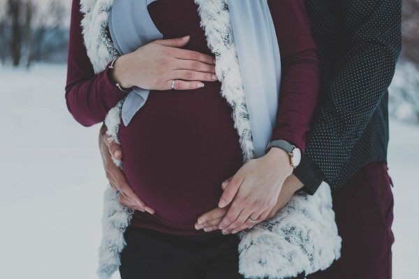 Der Bauch einer schwangeren Person ist im Fokus des Bildes. Sie tr&auml;gt ein dunkelrotes Kleid und eine helle Weste. Ihre H&auml;nde und die H&auml;nde einer weiteren Person liegen sch&uuml;tzend und liebevoll auf dem Babybauch. Die Szene spielt im Freien bei winterlicher Umgebung.