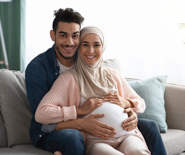 Ein lachendes Paar sitzt sich umarmend auf einem Sofa. Die schwangere Frau tr&auml;gt ein Kopftuch und h&auml;lt ihren Babybauch mit beiden H&auml;nden. Der Mann sitzt hinter ihr und legt ebenfalls seine H&auml;nde auf ihrem Bauch. Beide wirken gl&uuml;cklich und entspannt.