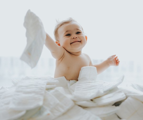 Ein Baby sitzt in einem Berg mit frischen wei&szlig;en Windeln und h&auml;lt eine davon in die Luft. Das Baby l&auml;chelt und sieht fr&ouml;hlich aus.