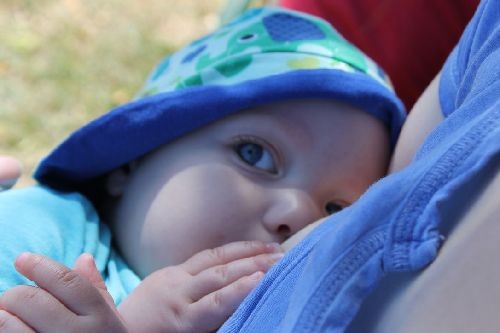 Ein Baby mit blau-gr&uuml;n gemustertem Hut liegt an der Brust einer stillenden Person. Es schaut mit gro&szlig;en Augen in die Kamera, w&auml;hrend es trinkt. Die Szene wirkt ruhig und innig.