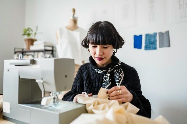 Eine Frau mit schwarzem Pullover und dunklen Haaren sitzt vor einer N&auml;hmaschine und h&auml;lt ein gro&szlig;es St&uuml;ck beigen Stoff in der Hand. Im Hintergrund steht eine Schneiderpuppe und ein Regal. 