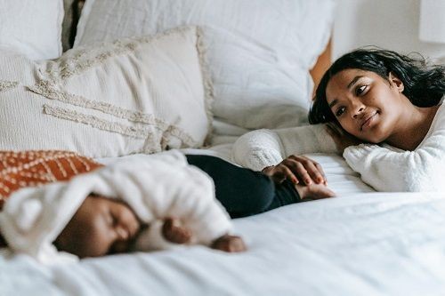 Ein Baby mit geschlossenen Augen und eine Frau liegen auf einem Bett. Die Frau l&auml;chelt das Baby an, hat ihren Kopf auf ihrem Arm abgelegt und h&auml;lt mit einer Hand den Fu&szlig; des Babys.