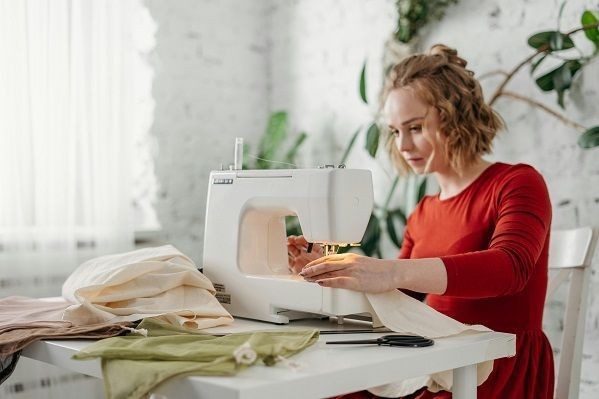 Eine Frau mit rotem Kleid und blondem Haaren n&auml;ht an einer N&auml;hmaschine, neben der Maschine liegt eine Schere und verschieden Stoffe. Im Hintergrund steht eine Pflanze.
