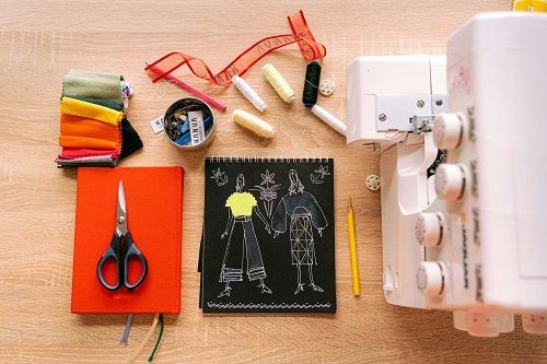 eine N&auml;hmaschine, 2 Bleistifte, ein Buch, ein Block, eine Schere, bunter Stoff, eine Dose mit Kn&ouml;pfen und Nadeln und ein paar Rollen Garn