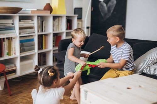 3 Kinder sind in einem Wohnzimmer und ziehen alle an einem Stofftierdrachen. Eines der Kinder h&auml;lt in der anderen Hand einen Kochl&ouml;ffel. Um die Kinder herum sind eine Couch, ein Kiste und ein tiefes B&uuml;cherregal zu sehen. 