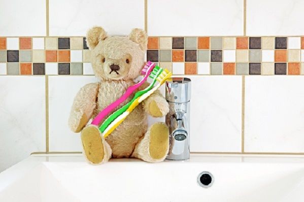 Ein Teddyb&auml;re sitzt neben einem Wasserhahn auf der Ablagefl&auml;che eines Waschbeckens und h&auml;lt drei verschiedenfarbige Zahnb&uuml;rsten im Arm.  