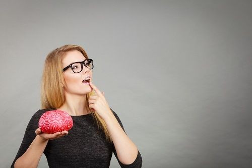 Eine Frau mit langen Haaren und Brille h&auml;lt eine Gehirn aus Plastik in der einen Hand und f&uuml;hrt den Zeigefinger der anderen Hand an die Unterlippe und schaut dabei nach oben.