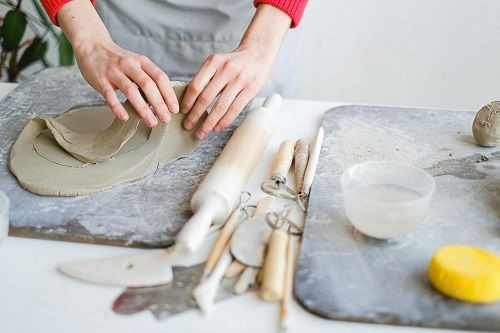 H&auml;nde schneiden einen Kreis aus ausgerolltem Ton heraus. Neben der Platte auf der gearbeitet wird, liegt verschiedenes T&ouml;pferwerkzeug und eine Teigwalze sowie ein Schwamm und eine Sch&uuml;ssel mit Wasser
