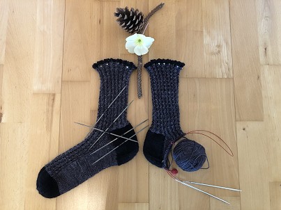 gestrickte Socken mit mehreren Stricknadeln und  einem Wollkn&auml;uel, &uuml;ber den Socken liegt ein Tannenzapfen, ein Ast und eine Blume 