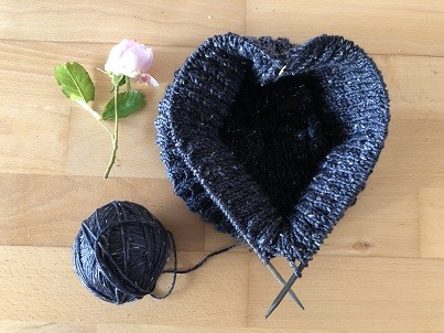 Ein Strickst&uuml;ck in dem 2 Stricknadeln stecken, die das Strickst&uuml;ck in eine Herzform f&uuml;hren. Neben dem Strickst&uuml;ck liegt eine Rose in rosa und der Rest der blauen Wolle.