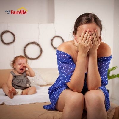 Eine Frau in einem blauen Kleid sitzt traurig auf einem Bett. Sie hat die H&auml;nde vor ihrem Gesicht. Im Hintergrund sitzt ein Baby und weint. Die Stimmung wirkt angespannt.