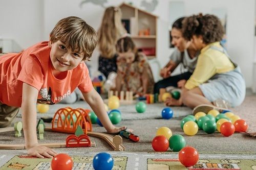 Ein Kind spielt im einer Eisenbahn auf Schienen, um es herum kleine bunte Plastikb&auml;lle. Im Hintergrund ist eine Frau mit mehreren Kindern zu sehen die mit Holzbausteinen, einem Spielhaus und anderen Dingen spielen. 
