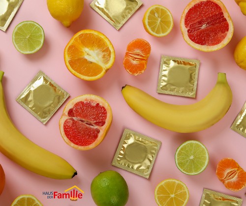 Auf einem rosa Untergrund sind mehrere Bananen, aufgeschnittene Orangen, Zitronen, Limetten und eine gesch&auml;lte, halbierte Mandarine zu sehen sowie mehrere Kondome und goldener Verpackung.