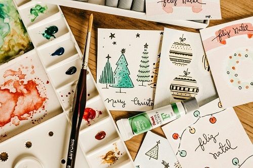 verschieden gestaltete Weihnachtskarten mit Tannenb&auml;umen, Chrtistbaumkugeln, Adventskranz und der Beschriftung frohe Weihnachten auf spanisch und englisch sowie der Mischbereich eines Malkasten mit Pinsel und Farben