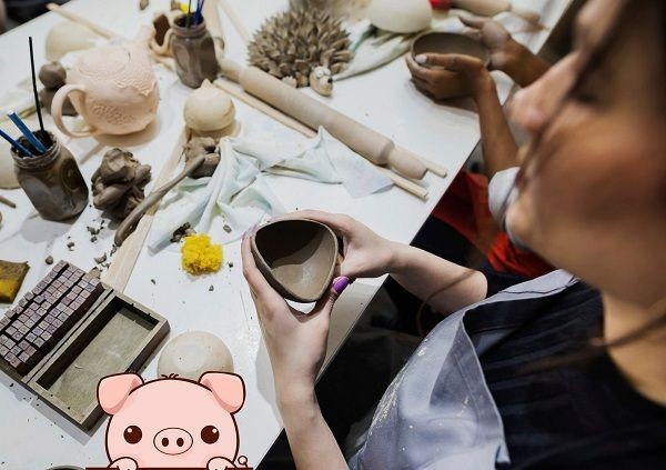 Tisch mit T&ouml;pfermaterial und Keramikst&uuml;cken an dem mehrere Personen arbeiten. Im Vordergrund w&uuml;rde ein Comicschwein eingef&uuml;gt, dass &uuml;ber den Bildrand schaut.
