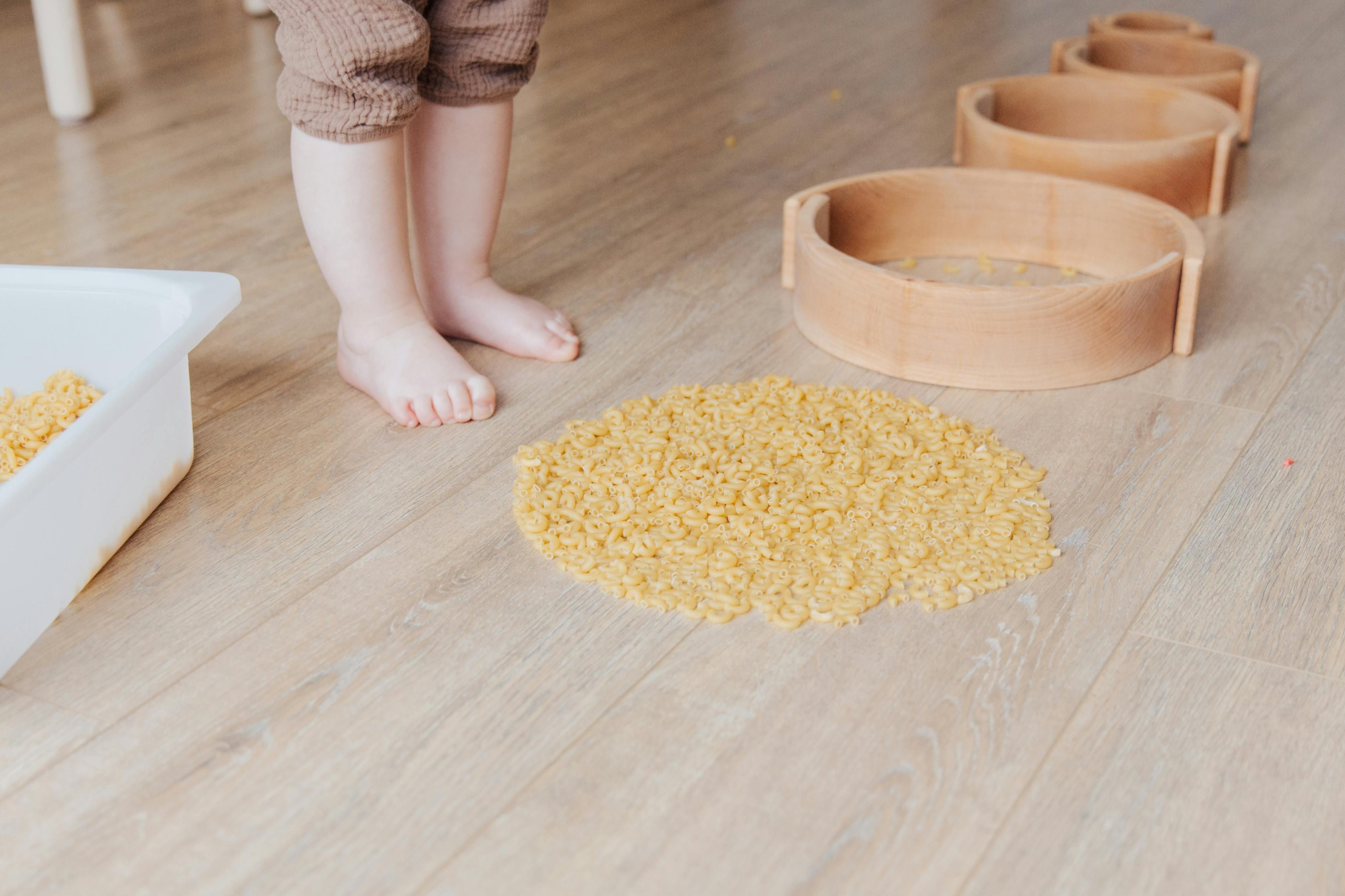 Ein Kind, von ihm sind nur die Beine und F&uuml;&szlig;e zu sehen, steht vor einem ausgef&uuml;llten Kreis aus Nudeln. Neben dem Kreis liegen 4 runde Holzformen, die von Form zu Form immer kleiner werden. Neben dem Kind liegt noch eine rechteckige Sch&uuml;ssel mit Nudeln. 