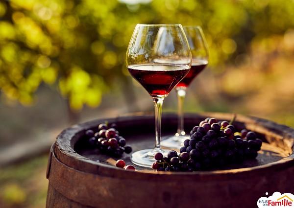 Zwei Gl&auml;ser Rotwein stehen auf einem Weinfass. Daneben liegen dunkle Weintrauben. Der Hintergrund ist gr&uuml;n durch B&auml;ume.
