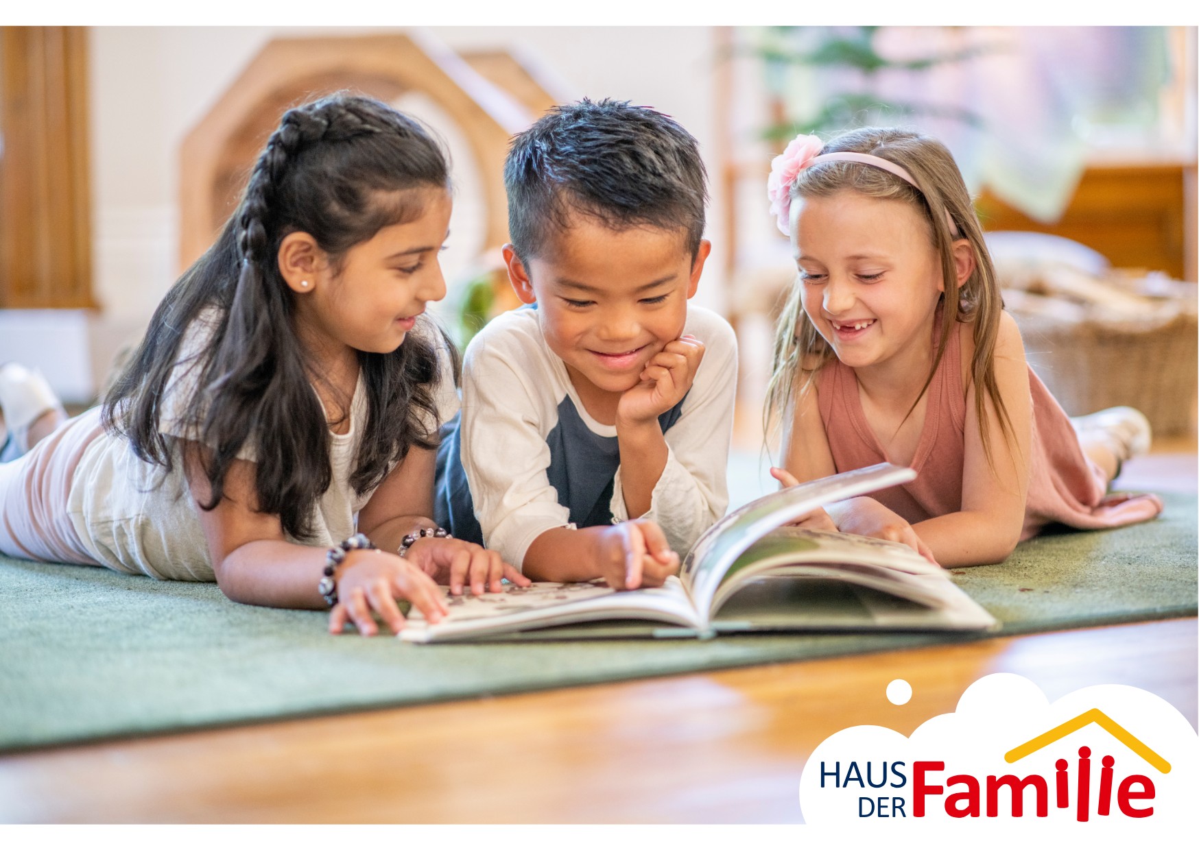Drei Kinder liegen auf einem Teppich und lesen gemeinsam ein Buch und l&auml;cheln.