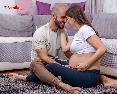 Ein werdendes Elternpaar sitzt sich l&auml;chelnd gegen&uuml;ber und h&auml;lt gemeinsam den Babybauch. Das Bild vermittelt N&auml;he, Vertrauen und gemeinsame Vorbereitung auf die Geburt.